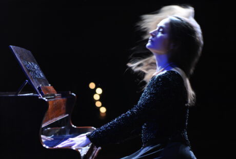 Brązowowłosa kobieta w czarnej sukni gra na fortepianie.